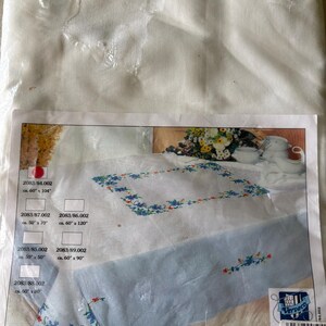 Op de afbeelding: Een verpakte witte tafelkleed met een bloemenborduurontwerp. De verpakking toont een tafelkleed met blauwe bloemenborduurwerk en afmetingen vari&euml;rend van 127cm x 127cm tot 152cm x 305cm. De merknaam "Vervaco" is zichtbaar.