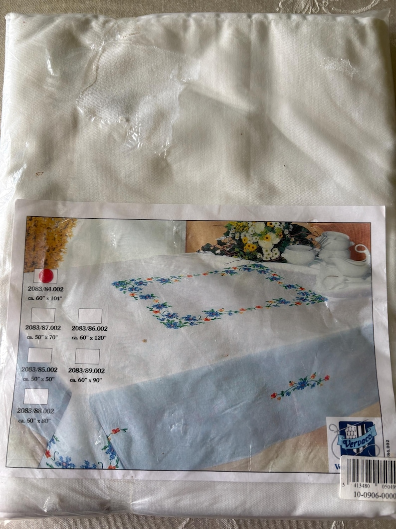 Op de afbeelding: Een verpakte witte tafelkleed met een bloemenborduurontwerp. De verpakking toont een tafelkleed met blauwe bloemenborduurwerk en afmetingen vari&euml;rend van 127cm x 127cm tot 152cm x 305cm. De merknaam "Vervaco" is zichtbaar.