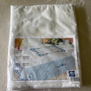 Op de afbeelding: Een verpakte witte tafelkleed met een bloemenontwerp. Het product is verzegeld in helder plastic. Het productlabel toont een tafelkleed met blauwe bloemenborduursels en verschillende maatopties.