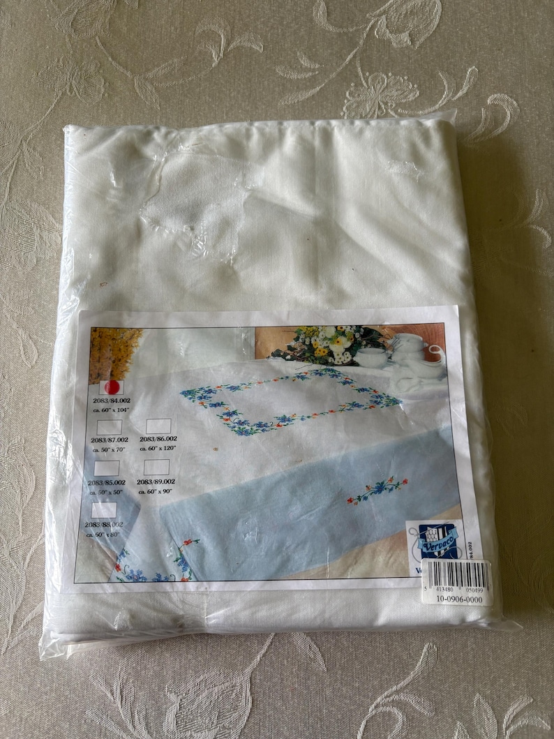 Op de afbeelding: Een verpakte witte tafelkleed met een bloemenontwerp. Het product is verzegeld in helder plastic. Het productlabel toont een tafelkleed met blauwe bloemenborduursels en verschillende maatopties.
