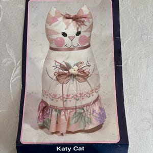 May include: A handmade Katy Cat doorstop. The cat-shaped doorstop features a white body with pink accents, a pink bow, and a floral skirt. The doorstop is made of fabric and has a cross-stitch design.