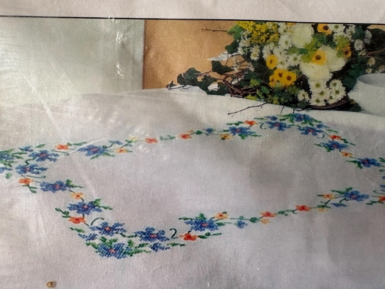 Op de afbeelding: Wit tafelkleed met een geborduurde bloemenrand in blauw, oranje en groen. Op de achtergrond staat een boeket witte en gele bloemen. De geborduurde rand vormt een rechthoek.