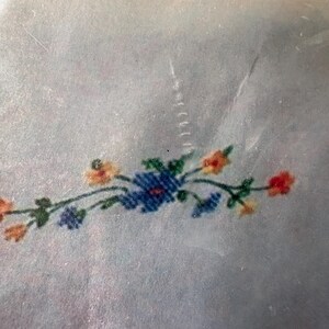 Op de afbeelding: Een close-up van een bloemenborduurontwerp. Het ontwerp toont een cluster van blauwe bloemen met oranje en rode accenten en groene bladeren. De borduursel is op een lichtblauwe stof.
