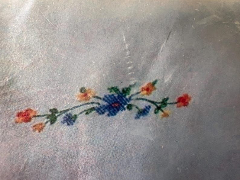 Op de afbeelding: Een close-up van een bloemenborduurontwerp. Het ontwerp toont een cluster van blauwe bloemen met oranje en rode accenten en groene bladeren. De borduursel is op een lichtblauwe stof.