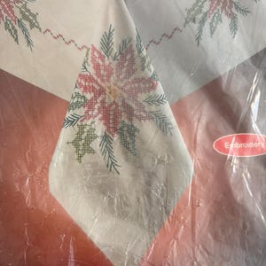 K&ouml;nnte beinhalten: Ein wei&szlig;es Tischtuch mit einem Kreuzstich-Poinsettien-Design in Rot, Gr&uuml;n und Wei&szlig;. Das Tischtuch ist in klarem Kunststoff verpackt. Das Wort "Embroidery" ist auf einem Aufkleber zu sehen.