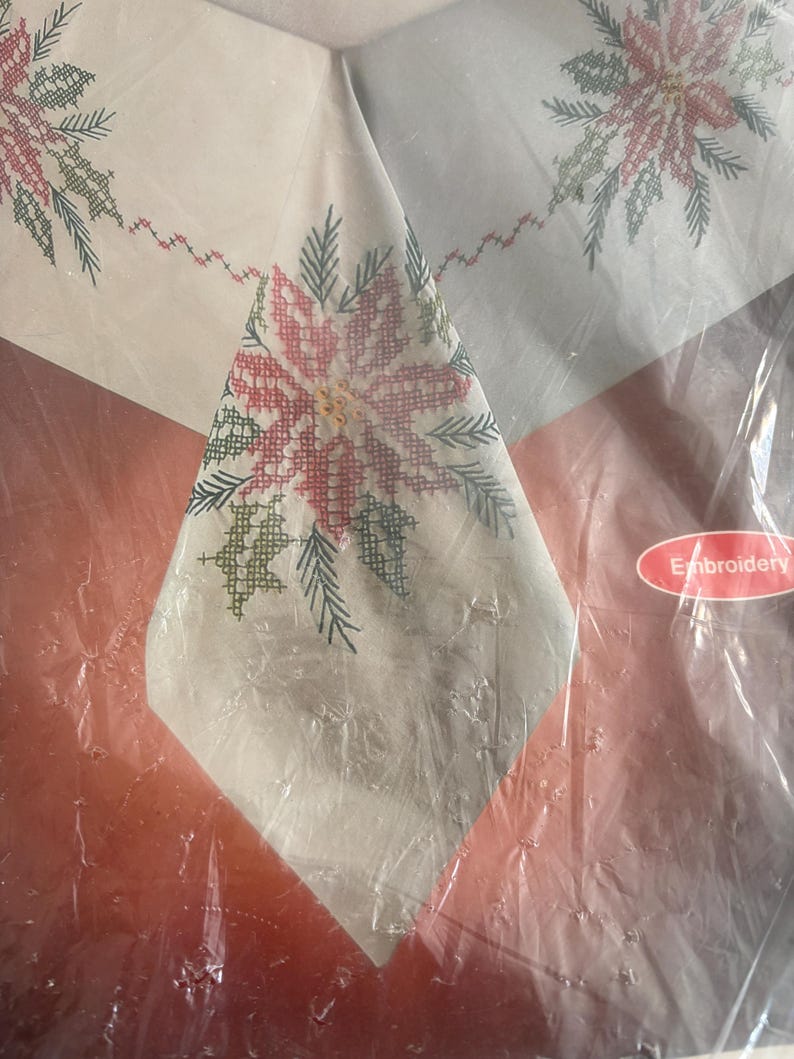 K&ouml;nnte beinhalten: Ein wei&szlig;es Tischtuch mit einem Kreuzstich-Poinsettien-Design in Rot, Gr&uuml;n und Wei&szlig;. Das Tischtuch ist in klarem Kunststoff verpackt. Das Wort "Embroidery" ist auf einem Aufkleber zu sehen.