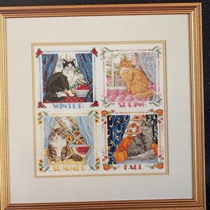 May include: Framed cross-stitch artwork featuring four panels, each depicting a cat in a seasonal setting. The panels are labeled "Winter," "Spring," "Summer," and "Fall." The frame is gold-colored.