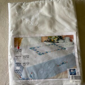 Op de afbeelding: Een verpakte witte tafelkleed met een bloemenontwerp. Het tafelkleed zit in een doorzichtige plastic verpakking. De productafbeelding toont een wit tafelkleed met een blauwe bloemenrand en een lichtblauw midden.