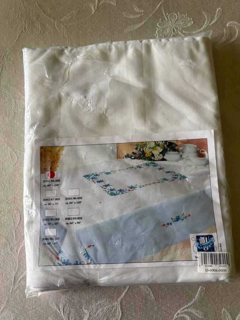 Op de afbeelding: Een verpakte witte tafelkleed met een bloemenontwerp. Het tafelkleed zit in een doorzichtige plastic verpakking. De productafbeelding toont een wit tafelkleed met een blauwe bloemenrand en een lichtblauw midden.