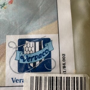 Op de afbeelding: Een close-up van een productlabel met de merknaam "Vervaco" in een blauw en wit logo. Het label bevat een barcode en het nummer "5 413480 050499". De achtergrond toont een lichtblauwe stof met bloemenborduurwerk.