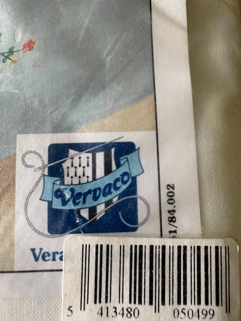 Op de afbeelding: Een close-up van een productlabel met de merknaam "Vervaco" in een blauw en wit logo. Het label bevat een barcode en het nummer "5 413480 050499". De achtergrond toont een lichtblauwe stof met bloemenborduurwerk.