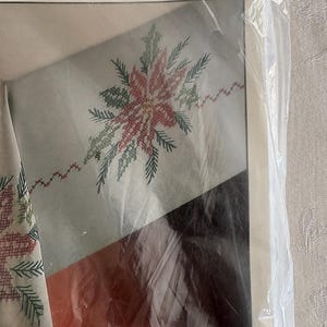 K&ouml;nnte beinhalten: Ein verpacktes Stickset mit einem Poinsettien-Design. Die Tischdecke besteht aus einer Baumwollmischung und hat eine schmutzabweisende Oberfl&auml;che. Das Set enth&auml;lt Stickgarn und Anleitungen. Auf der Verpackung steht das Wort "Stickerei".