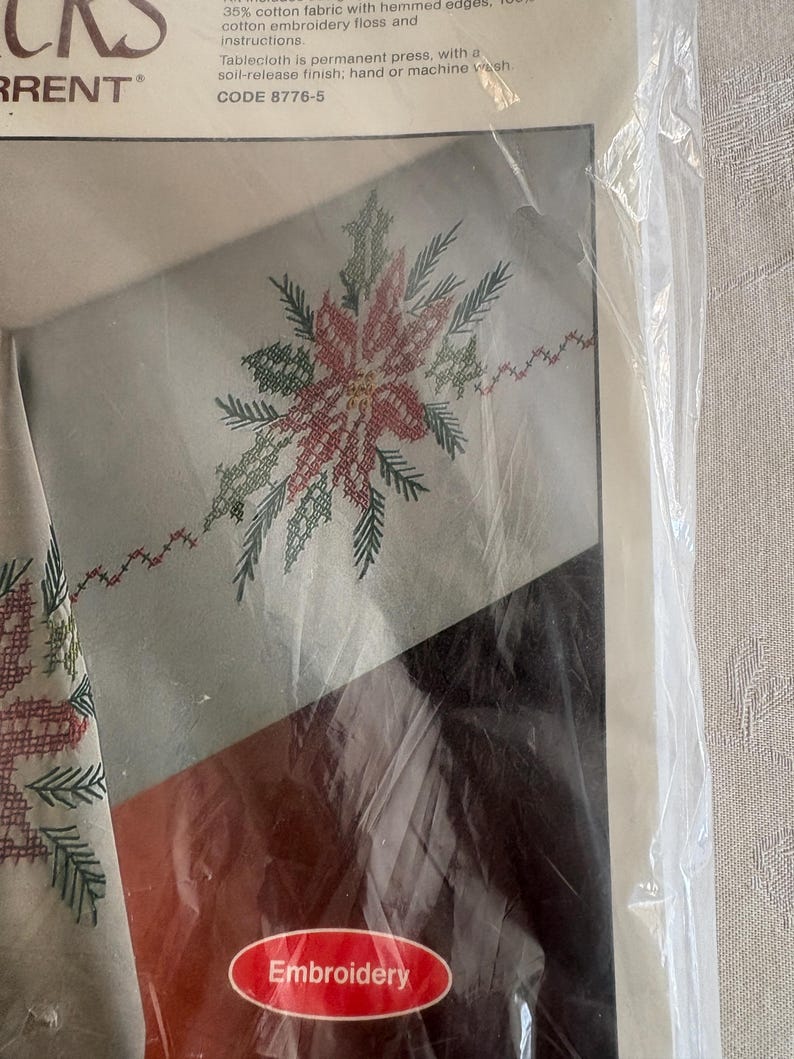 K&ouml;nnte beinhalten: Ein verpacktes Stickset mit einem Poinsettien-Design. Die Tischdecke besteht aus einer Baumwollmischung und hat eine schmutzabweisende Oberfl&auml;che. Das Set enth&auml;lt Stickgarn und Anleitungen. Auf der Verpackung steht das Wort "Stickerei".