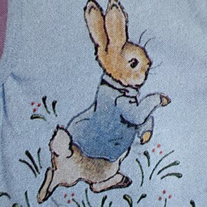 May include: A light blue fabric featuring a printed illustration of a brown rabbit wearing a blue jacket. The rabbit is depicted in a running pose amidst small green grass blades and tiny red floral accents, creating a repeating pattern on the textile.