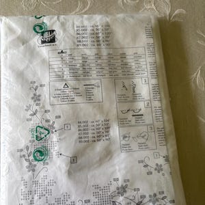 Op de afbeelding: Een pakket met een DMC borduurpatroon. Het patroon bevat bloemmotieven en tekst met afmetingen in centimeters. Het pakket is verzegeld in helder plastic en heeft een groen recycling symbool.