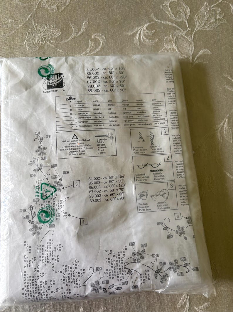 Op de afbeelding: Een pakket met een DMC borduurpatroon. Het patroon bevat bloemmotieven en tekst met afmetingen in centimeters. Het pakket is verzegeld in helder plastic en heeft een groen recycling symbool.