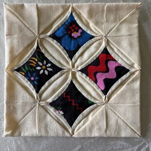 May include: A square, handmade fabric coaster with a cream-colored background. The coaster features a diamond pattern with four colorful floral and abstract designs in the center. The designs include blue, red, and yellow floral patterns.