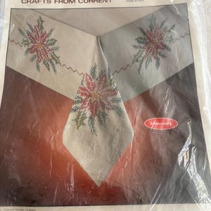 K&ouml;nnte beinhalten: Ein verpacktes Fiddlesticks-Bastelset mit einer bestickten Tischdecke mit einem Holiday-Spray-Design. Die Tischdecke hat einen cremefarbenen Hintergrund mit roter und gr&uuml;ner Blumenstickerei. Der Text "Fiddlesticks CRAFTS FROM CURRENT" ist enthalten.
