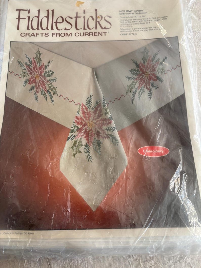 K&ouml;nnte beinhalten: Ein verpacktes Fiddlesticks-Bastelset mit einer bestickten Tischdecke mit einem Holiday-Spray-Design. Die Tischdecke hat einen cremefarbenen Hintergrund mit roter und gr&uuml;ner Blumenstickerei. Der Text "Fiddlesticks CRAFTS FROM CURRENT" ist enthalten.