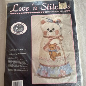 May include: A cross-stitch kit titled "Love 'n Stitches" featuring a design of a bear named "Bindy Bear." The kit includes cotton embroidery floss, Aida cloth, ribbon, and a pattern. Finished size is 10" x 14".