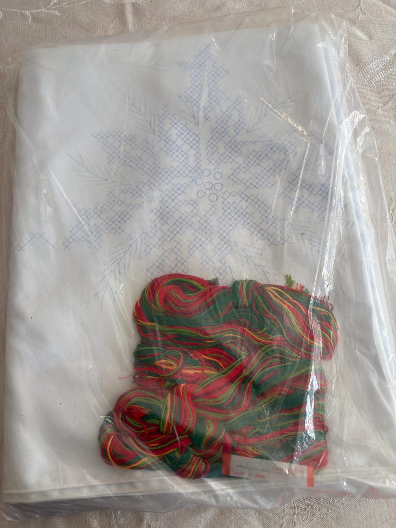 K&ouml;nnte beinhalten: Ein wei&szlig;er Stoff mit blauem Schneeflockenmuster, verpackt in durchsichtigem Kunststoff.  Mit rotem, gr&uuml;nem und gelbem Stickgarn. Ein Bastelset f&uuml;r Nadelmalerei oder Kreuzstich.