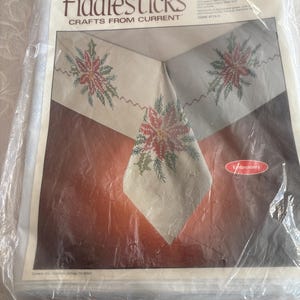 K&ouml;nnte beinhalten: Ein verpacktes Stickset von Fiddlesticks mit dem Design Holiday Spray. Das Set enth&auml;lt einen cremefarbenen Stoff mit gestickten Weihnachtssternen und gr&uuml;nem Laub. Die Verpackung ist in klarem Kunststoff versiegelt.