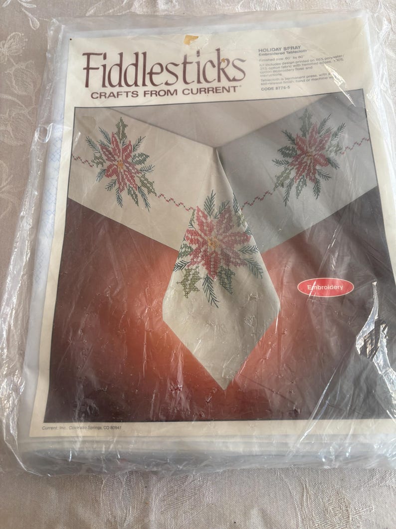 K&ouml;nnte beinhalten: Ein verpacktes Stickset von Fiddlesticks mit dem Design Holiday Spray. Das Set enth&auml;lt einen cremefarbenen Stoff mit gestickten Weihnachtssternen und gr&uuml;nem Laub. Die Verpackung ist in klarem Kunststoff versiegelt.