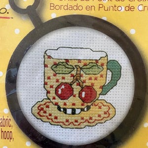 May include: A counted cross stitch kit featuring a yellow teacup with a red cherry pattern, a green handle, and a scalloped saucer. The design is displayed inside a black circular embroidery hoop. The packaging includes the text Counted Cross Stitch.