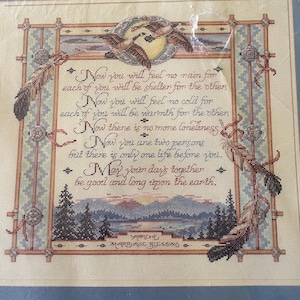 May include: A framed cross-stitch sampler featuring the Apache Marriage Blessing text in brown script. The design includes two geese in flight, feathers, and a landscape with mountains and pine trees in muted earth tones, all enclosed in a wooden frame.