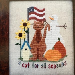 May include: A cross-stitch embroidery on light fabric featuring a brown cat standing in the center. The design includes a waving American flag, two yellow sunflowers, a blue-roofed birdhouse, a white snowman, and two orange pumpkins. The text "a cat for all seasons" is stitched in dark letters at the bottom.