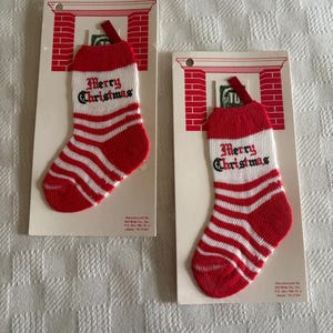 May include: Two miniature knit Christmas stockings on individual cardboard display cards. Each stocking features red and white horizontal stripes, a red cuff, and the text Merry Christmas in black script. The cards show a red brick fireplace graphic.