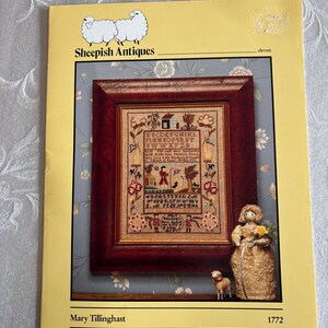 May include: A vintage cross-stitch pattern booklet titled "Sheepish Antiques" featuring a framed sampler with a detailed design. The booklet includes a small figurine of a woman and a dog. The cover is yellow.