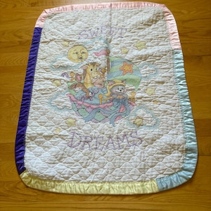 May include: A pastel-coloured baby blanket with a quilted pattern. The blanket features a central design of cartoon animals on a boat, with the words "SWEET DREAMS" embroidered on it. The edges are trimmed with satin in various colours.