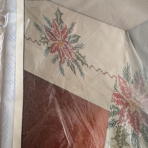 K&ouml;nnte beinhalten: Ein Kreuzstich-Bastelset mit einem Poinsettien-Design. Das Set enth&auml;lt einen cremefarbenen Stoff mit einem detaillierten Muster aus roten, gr&uuml;nen und gelben Blumen und Bl&auml;ttern. Der Text "Crafts From Current" ist oben auf der Verpackung zu sehen.