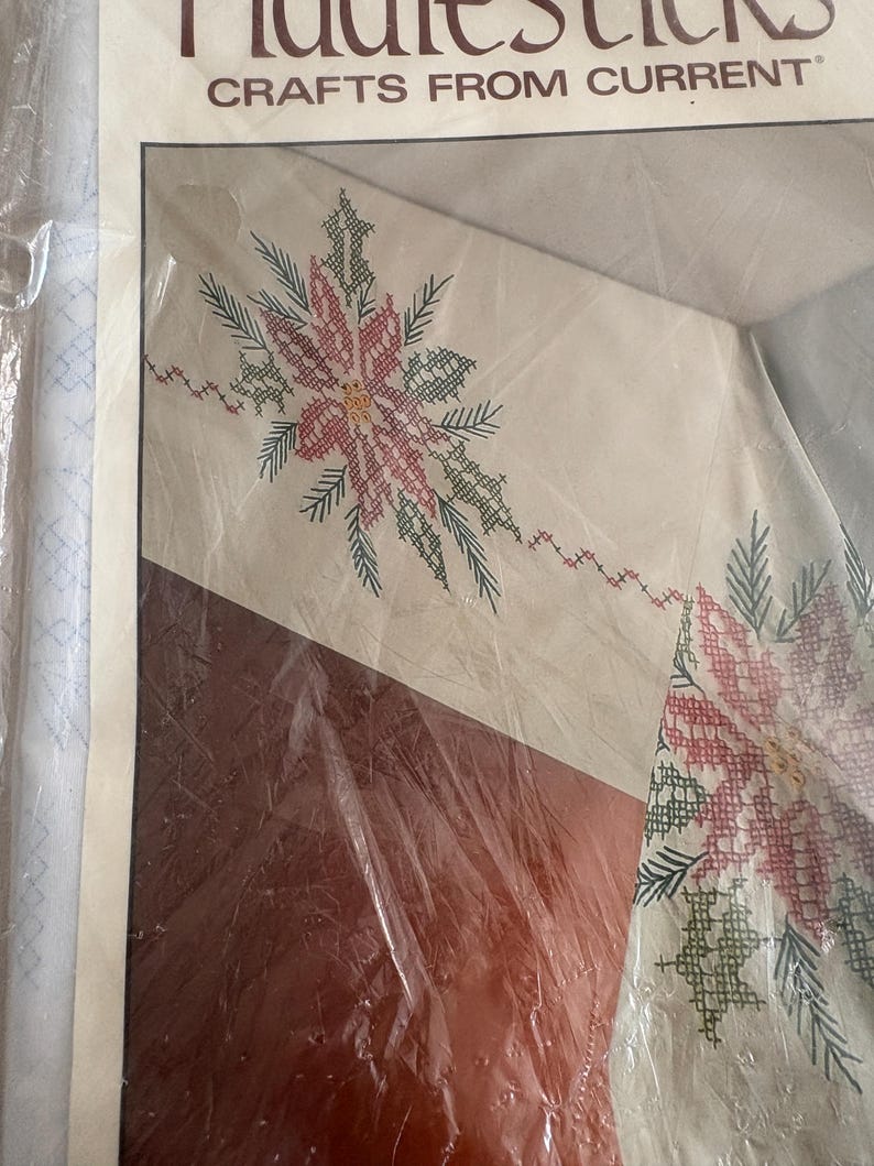 K&ouml;nnte beinhalten: Ein Kreuzstich-Bastelset mit einem Poinsettien-Design. Das Set enth&auml;lt einen cremefarbenen Stoff mit einem detaillierten Muster aus roten, gr&uuml;nen und gelben Blumen und Bl&auml;ttern. Der Text "Crafts From Current" ist oben auf der Verpackung zu sehen.