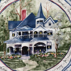 May include: A fabric panel featuring a detailed illustration of a Victorian-style house with a blue roof and white exterior. The house is surrounded by trees and flowers, with a path leading to the front door. The panel has a circular border with text.