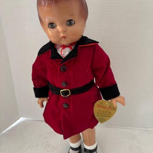May include: A vintage Patsy Ann doll wearing a red coat with black trim and a black belt with a gold buckle. The doll has black shoes, white socks, and a tag that reads "The Patsy Ann".
