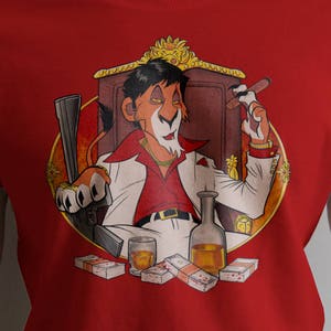 May include: Red t-shirt with a cartoon illustration of a lion wearing a red shirt, gold chain, and a crown. The lion is holding a glass of liquor and a cigar. The lion is sitting on a throne with money scattered around him.