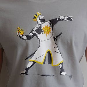 May include: A gray t-shirt with a graphic print of a knight in white armor holding a golden orb. The knight is wearing a crown and has a yellow sun on his chest. The graphic is in a spray paint style.