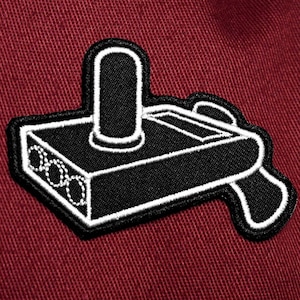 Peut inclure: Un patch brodé noir et blanc représentant un joystick de jeu vidéo vintage. Le joystick est sur un fond en tissu rouge.