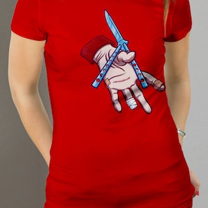 Puede incluir: Camiseta roja con una ilustraci&oacute;n de dibujos animados de una mano sosteniendo dos cuchillos mariposa azules.