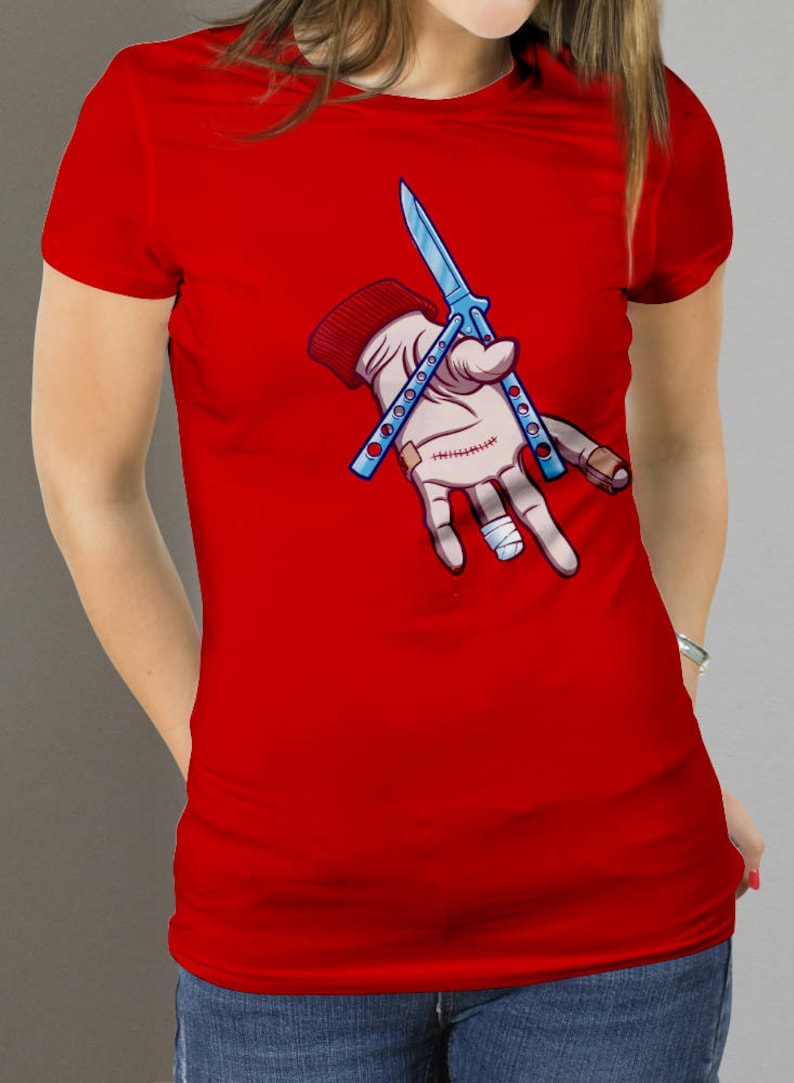 Puede incluir: Camiseta roja con una ilustraci&oacute;n de dibujos animados de una mano sosteniendo dos cuchillos mariposa azules.