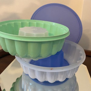 May include: Three plastic, stackable, round, clear, and colored jello molds. The top mold is green, the middle mold is blue, and the bottom mold is clear with a blue base. The top mold has a matching lid that is light blue.