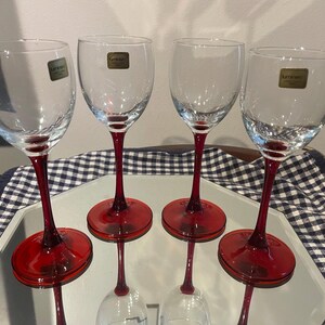 May include: A set of four clear glass wine glasses with deep red stems and circular bases. Each glass features a small, rectangular gold-colored sticker on the bowl with the text Luminarc printed in black, indicating the brand of the glassware.