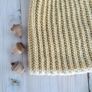 May include: A close-up of a beige knitted hat with a white trim. The hat is lying on a white wooden surface with three acorns beside it.