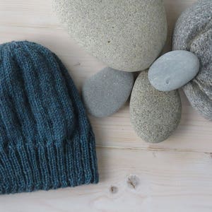 Puede incluir: Un gorro de punto azul oscuro con textura acanalada. El gorro est&aacute; sobre una superficie de madera clara con varias piedras grises lisas.