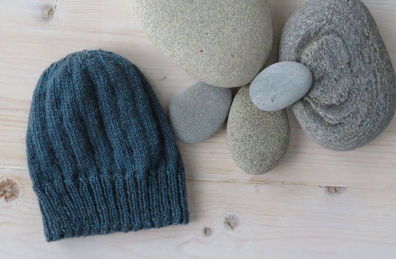 Puede incluir: Un gorro de punto azul oscuro con textura acanalada. El gorro est&aacute; sobre una superficie de madera clara con varias piedras grises lisas.