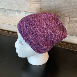 May include: A purple knitted beanie hat with a textured pattern. The hat is displayed on a white mannequin head.