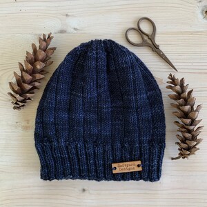 Puede incluir: Un gorro de punto azul oscuro con una etiqueta de cuero marr&oacute;n que dice "Soft yarn Designs".