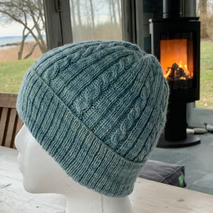 Peut inclure: Un bonnet en tricot bleu sarcelle avec un motif en torsades. Le bonnet est sur une tête de mannequin blanche.